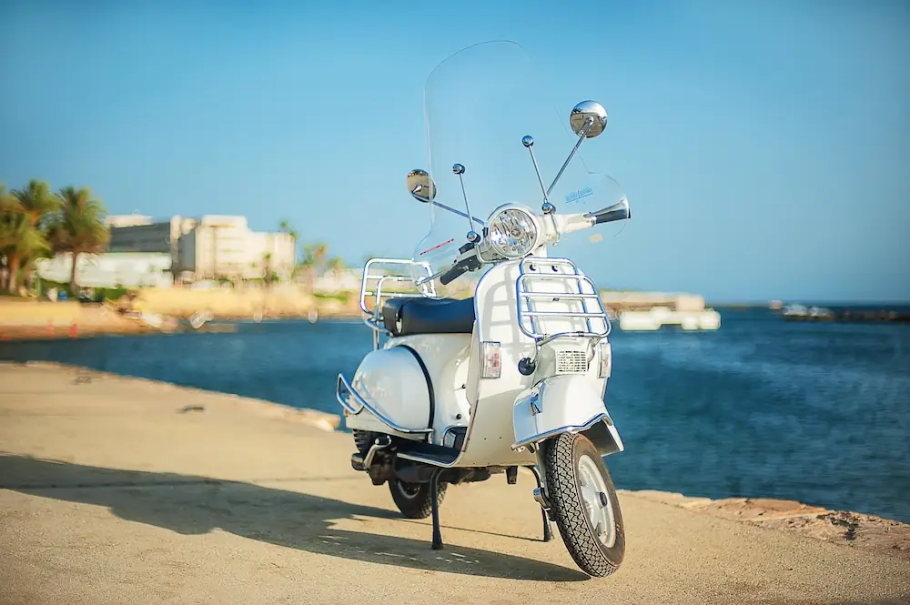 Witte scooter geparkeerd aan de kust met blauwe lucht en water op de achtergrond.