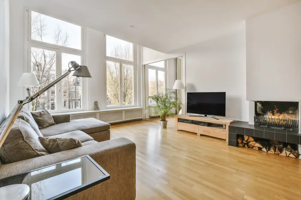 Een lichte woonkamer met grote ramen, een hoekbank, televisie en open haard.