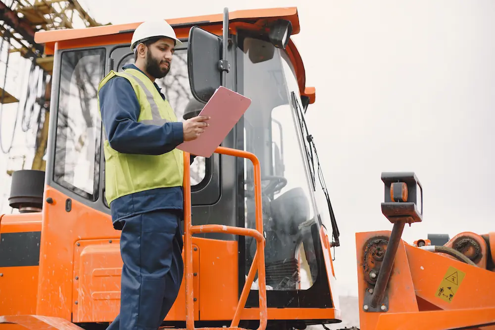 Een man met een veiligheidshelm en -vest inspecteert een stuk oranje werkmaterieel met een clipboard.