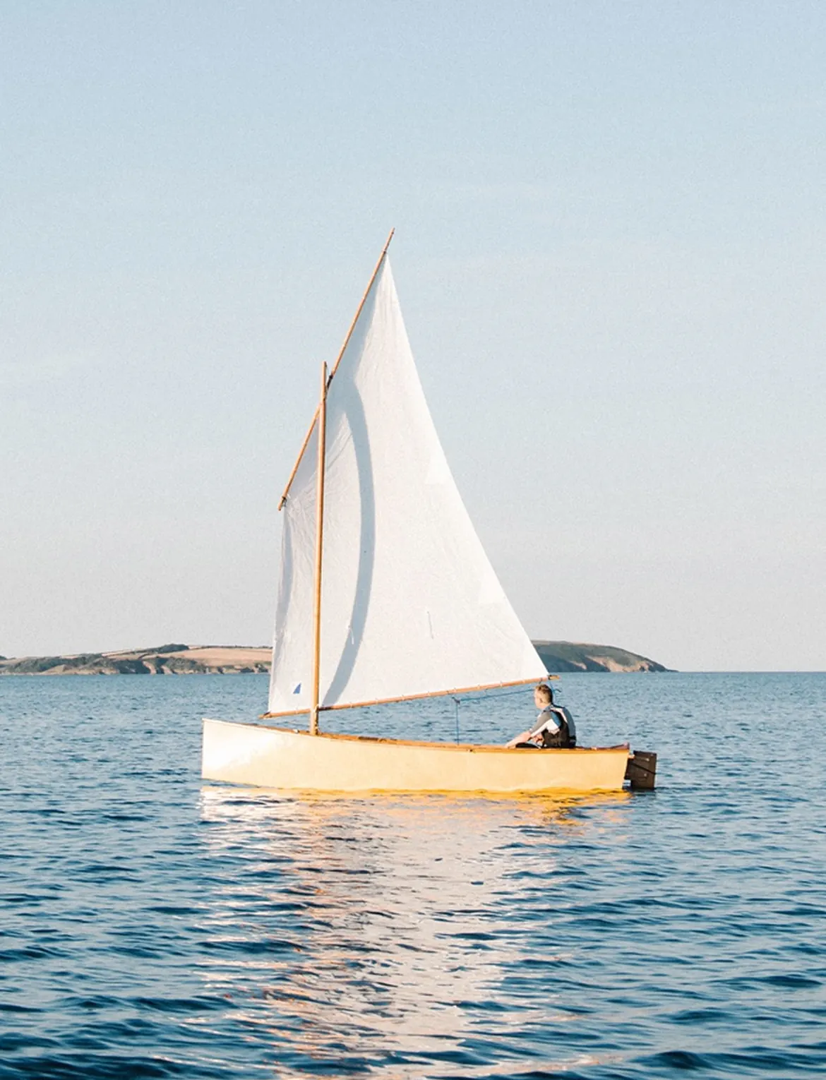 Een persoon zeilt in een kleine, gele zeilboot op een rustige zee met een helderblauwe lucht op de achtergrond.
