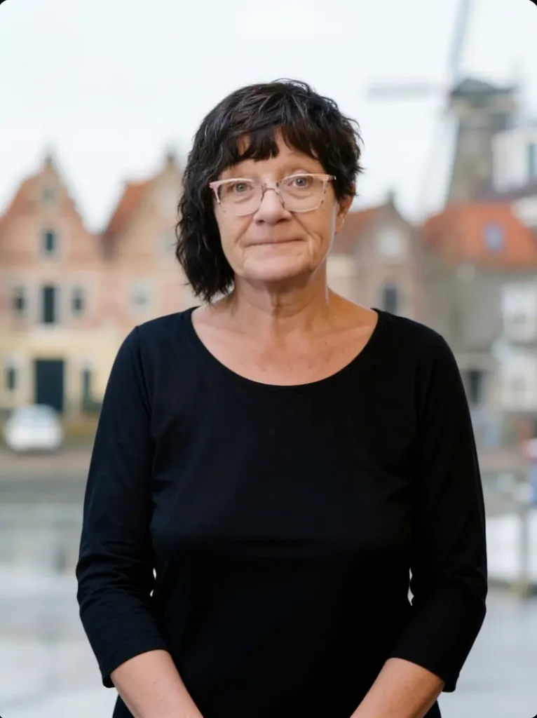 Een vrouw met donker krullend haar, een zwarte top en een bril staat voor een achtergrond met traditionele Nederlandse huisjes en een molen.