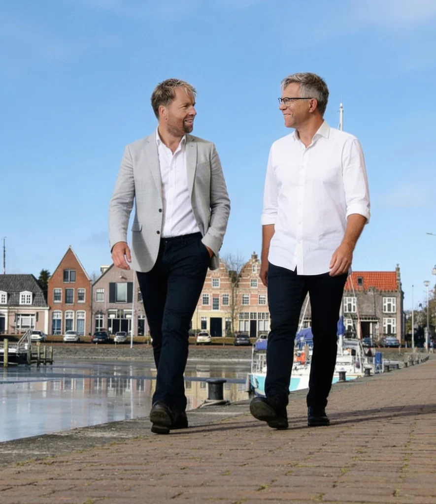 Twee mannen wandelen naast een haven met traditionele huizen op de achtergrond.