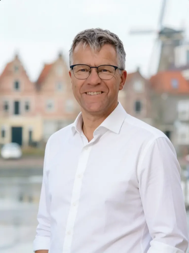 Een man met een bril en wit overhemd poseert met een Nederlandse achtergrond van klassieke gebouwen en een windmolen.