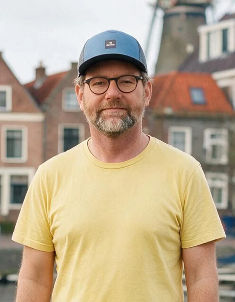 Een persoon met een baard en bril draagt een geel T-shirt en een blauwe pet, met traditionele Europese huizen op de achtergrond.