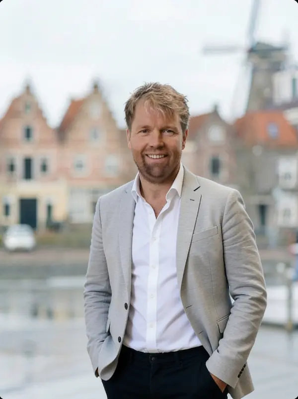 Man in een grijs pak poseert glimlachend voor traditionele Nederlandse huizen met een windmolen op de achtergrond.