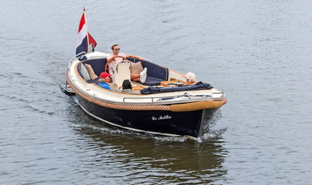 Een sloep genaamd "De MelRa" vaart met mensen aan boord op een rustige waterweg.